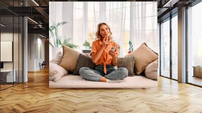 Technology and lifestyle. Attractive woman touching chin and looking thoughtfully aside while sitting on sofa with cell phone. Blonde female with crossed legs resting at home and using modern gadget. Wall mural