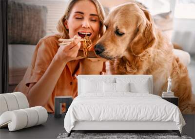 Togetherness with animal. Golden retriever eagerly licking paper box with chinese noodles being eating by young woman. Female pet owner sharing food with lovely furry friend at cozy apartment. Wall mural