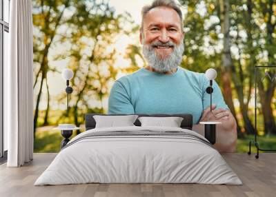 Vitality and active lifestyle. Portrait of fit mature man with hands crossed posing on camera during active training in green forest. Grey-bearded sportsman in blue shirt making confident smile. Wall mural