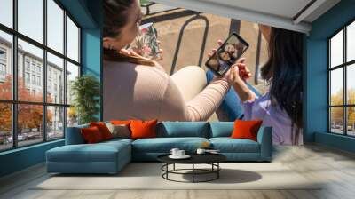 Two girl friends sitting on a bench showing each other a photo on their cell phone. Technology and communication concept Wall mural