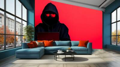 A mysterious figure in a hooded jacket working on a laptop against a bold red background, symbolizing anonymity and cyberspace. Wall mural