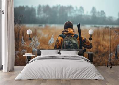 Hunter in a field with a gun and a backpack A man in camouflage clothing with a backpack and a gun walks through a field of tall grass Wall mural