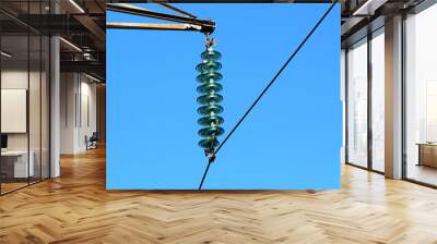 Layered high quality tall glass power line utility insulators holding single black electrical wire on strong metal utility pole on clear blue sky background Wall mural