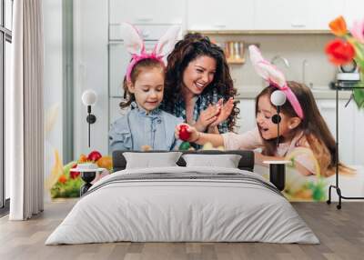 A mother and her cute daughters are painting eggs. Happy family are preparing for Easter. They are all wearing bunny ears. Wall mural