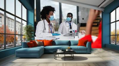 Afro American doctors talking with their patient behind protective glass. They are wearing protective face masks as a virus pandemic protection. Coronavirus healthcare concept. Wall mural