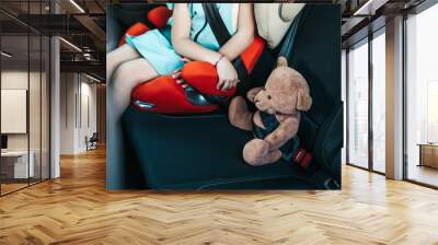 Little girl sitting in a car on a safety child car seat with her teddy bear. Wall mural