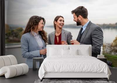 Three business people standing on the roof of modern building. They are drinking coffee, talking and smiling. Beautiful river landscape in the background. Wall mural