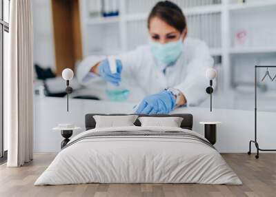 Young female practitioner or nurse with face protective mask working at clinic reception desk. She is holding disinfectant bottle and cleaining. Selective focus on hand. Wall mural