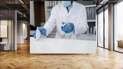 Young male practitioner with face protective mask working at clinic reception desk. He is holding disinfectant bottle and disinfect desk surface. Wall mural