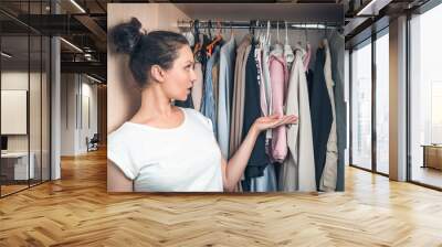 The woman looks at the things hanging in the wardrobe in surprise and thoughtfully. The concept of home life. Wall mural