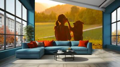 CLOSE UP: Adorable shot of a puppy putting its paw on cheerful woman meditating in the golden lit countryside. Beautiful view of a young woman relaxing in nature and bonding with her dog at sunset. Wall mural