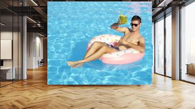 CLOSE UP Happy young guy on summer vacation lying on inflatable doughnut floatie in swimming pool & drinking pineapple alcohol beverage. Smiling man floating on inflatable pillow and sipping cocktail Wall mural