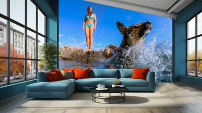 LOW ANGLE VIEW: Lady in bikini stands on sea rock when her dog jumps into water. Upon landing in crystal blue sea, adorable brown doggo makes a big splash. Active summer holidays at Dalmatian seaside. Wall mural