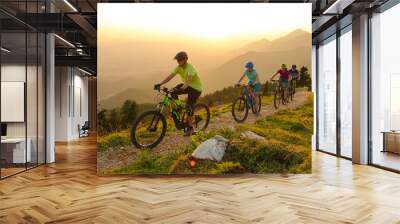 SUN FLARE Cheerful tourists ride electric bicycles up a mountain trail at sunset Wall mural