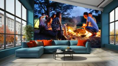 a company of friends have a rest in the evening on the banks of the river in nature. They fry sausages and talk. Cozy atmosphere, light from the fire. Summer rest, background Wall mural