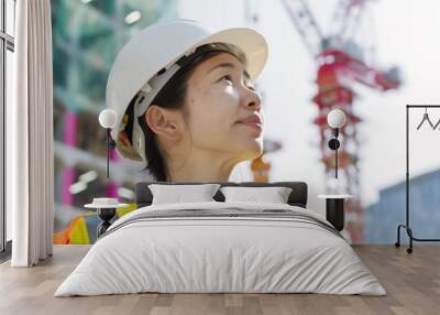 Asian female civil engineer in hard hat and safety vest on construction site examining high-rise framework with cranes in background Wall mural