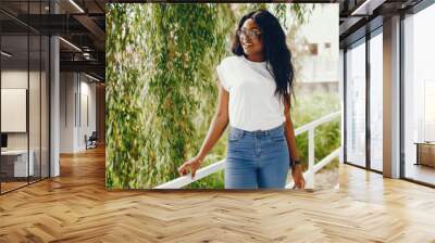 Cute black girl in a park. Lady in a white t-shirt and blue jeans. Woman in a sunglasses Wall mural