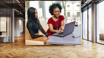 two beautiful and stylish young, dark girls sitting in a restaurant at the table, chatting and using a laptop Wall mural