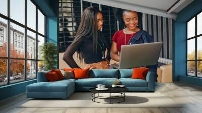 two beautiful and stylish young, dark girls standing in a restaurant and using a laptop Wall mural