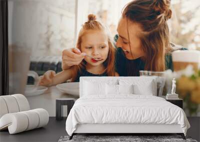 young and stylish mother with long hair and a green dress sitting with her little cute daughter in the summer cafe and she feeds her daughter with a dessert Wall mural