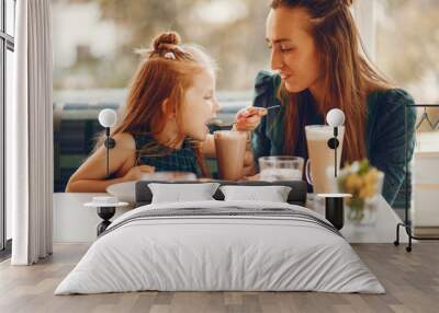 young and stylish mother with long hair and a green dress sitting with her little cute daughter in the summer cafe and she feeds her daughter with a dessert Wall mural