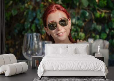 A beautiful young red-haired woman smiling while posing with a drink in her hand at a picnic table on a summer day outdoors Wall mural