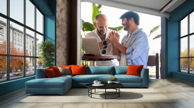 Two middle-aged male professionals are happily working on their laptops in the garden of a café on a summer day. Upon receiving some good news, they celebrate with joyful gestures and expressions Wall mural