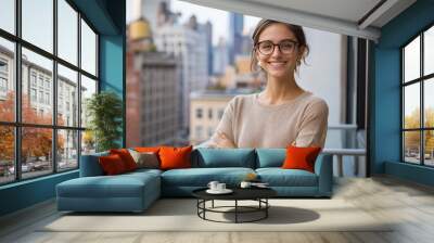 A confident woman stands on a balcony, her arms crossed and a radiant smile on her face. She wears stylish glasses and embodies pride in her role at the company. The urban landscape of New York serves Wall mural