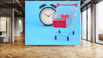 Shopping cart and classic alarm clock on blue background. Sale time buy mall market shop consumer concept. Selective focus. Wall mural