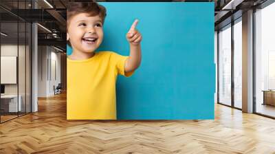 A cheerful boy in a yellow shirt is pointing and laughing on a blue background Space available for text Wall mural