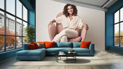 A millennial lady posing and relaxing indoors sitting in an armchair against a white studio wall smiling at the camera in a full length portrait Wall mural