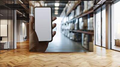 Hand holding a blank mobile phone in a blurred warehouse setting depicting online shopping mobile payments ordering products and business logistics with copy space image Wall mural