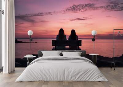 Two faceless girls sit on a bench overlooking calm water a rocky seashore and a pink sky at evening People communicate on the horizon while ships sail at sea. silhouette concept Wall mural