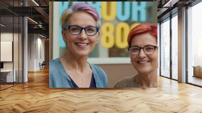 Two mature women, their vibrant attire mirroring the colorful art exhibition around them, share a moment of friendship. Their shared interest in art, evident in their engagement with the shop windows. Wall mural