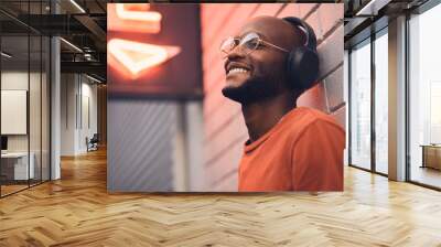 .Thinking, smile and music with a black man in the city, leaning on a brick wall on the street at night. Idea, glasses and headphones with a happy young male person streaming or listening to audio. Wall mural