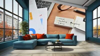 There is a piece of paper with a graph printed on it, a clipboard, and an open vocabulary book on the desk. There is the word Chief Marketing Officer on it. Wall mural