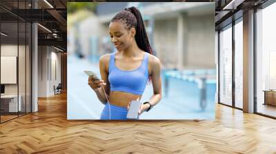 Beautiful and attractive sport woman in sport wear holding mobile phone and charger phone. Powerbank and smartphone in girl's hands. Energy charger power bank smart phone. Wall mural