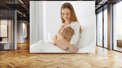 Beautiful mother breast-feeding her baby girl, sitting on bed and hugging little daughter, proper healthy breastfeeding of children Wall mural