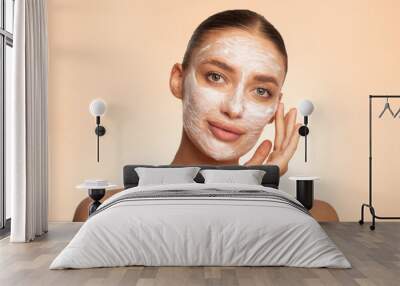 Beautiful young European woman applying facial mask on her face, enjoying skin care and treatment over beige background Wall mural