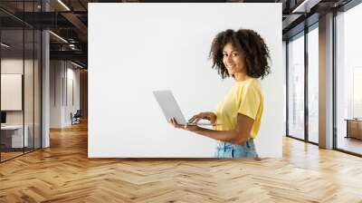 Cheerful black woman holding a laptop in her hands and pressing her finger on the keyboard while working online on a white wall background. Empty space Wall mural