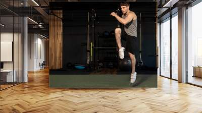 Energetic man doing fitness exercise, warming up and lifting leg, full length shot, gym interior Wall mural