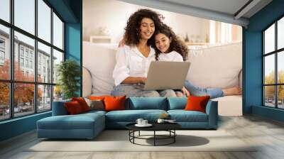 Family digital leisure. Cheerful latin mother and daughter using laptop engaging in fun online activities, surfing internet or video calling sitting on sofa at home, free space Wall mural