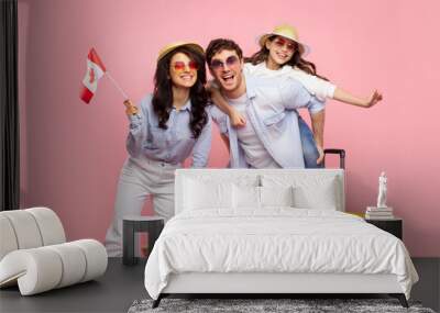 Family vacation. Happy family of three travelling together, woman holding Canada flag, posing with suitcase on pink background Wall mural