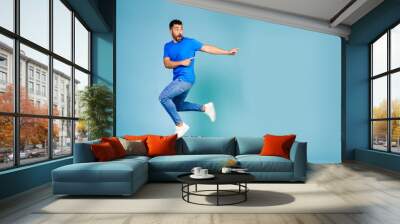 Full length portrait of cheerful young man jumping in blue t-shirt pointing finger to the side celebrating success against blue background Wall mural