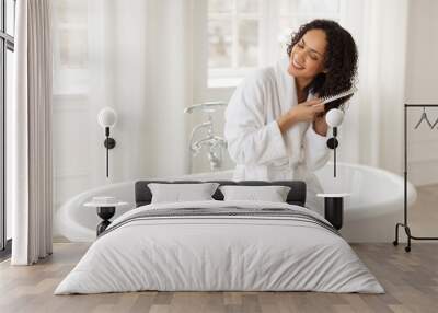 Haircare. African american woman brushing hair with comb, detangling split ends and smiling, sitting on bathtub in white bathrobe. Beauty routine concept Wall mural