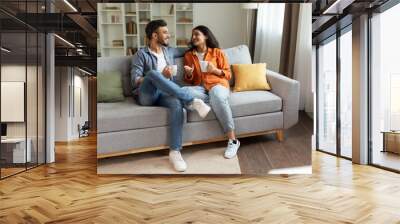 Happy indian couple relaxing and drinking coffee, sitting on couch at home, talking while resting with hot drinks in cups at modern living room, free space Wall mural