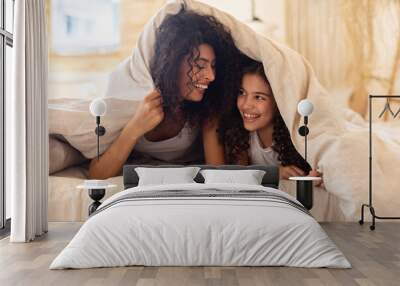 Happy joyful latin mother and adorable daughter hiding under soft comforter on comfortable bed, enjoying lazy morning and laughing, relaxing on fresh bed linen Wall mural