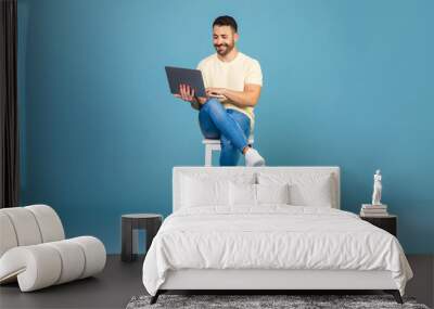 Happy man using laptop computer sitting on chair, working online over blue studio background, full length, copy space Wall mural