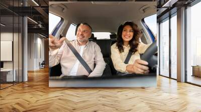 Happy middle aged man pointing with finger at the road and saying something while woman driving car, enjoying trip together Wall mural