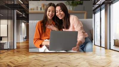 Happy mother and teen daughter looking at laptop, doing online shopping or watching movie using computer, sitting on sofa in living room Wall mural